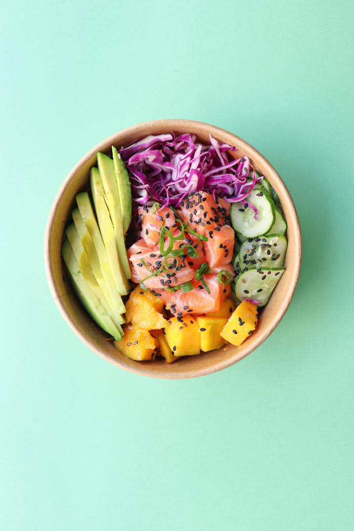 Top view of a vibrant poke bowl with salmon, avocado, mango, and vegetables on a mint background.